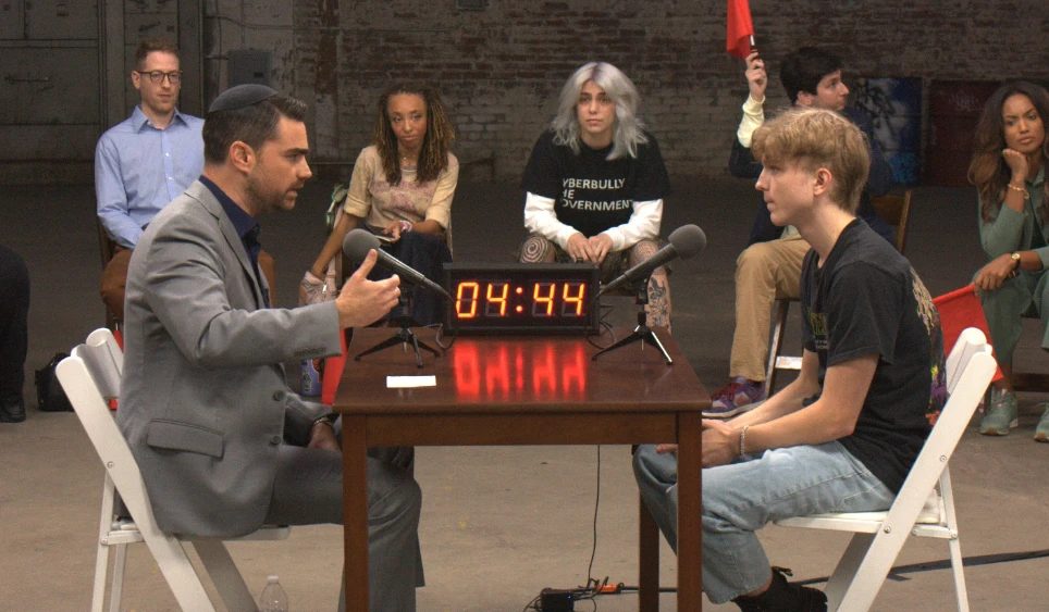 A one on one debate at a table with a countdown clock between the speakers and an audience seated around them