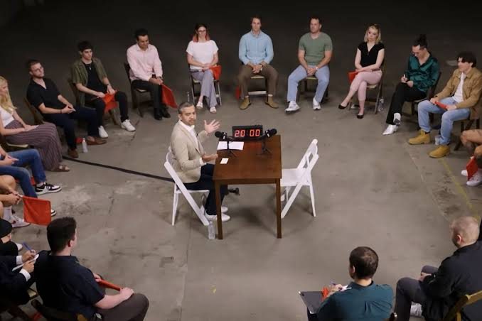 A speaker at a table in the middle of a circle of seated people holding coloured cards
